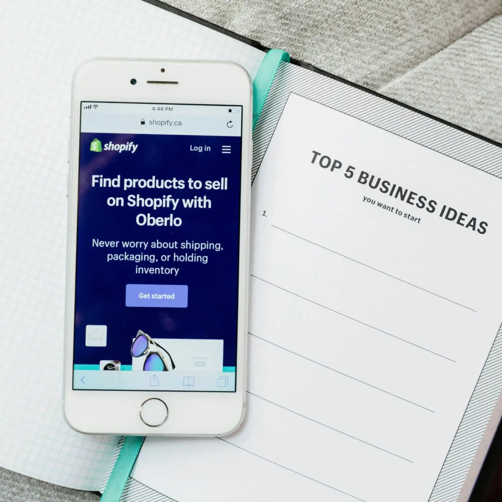 Smartphone displaying Shopify Oberlo page for finding winning shopify product to sell next to a notebook with business ideas