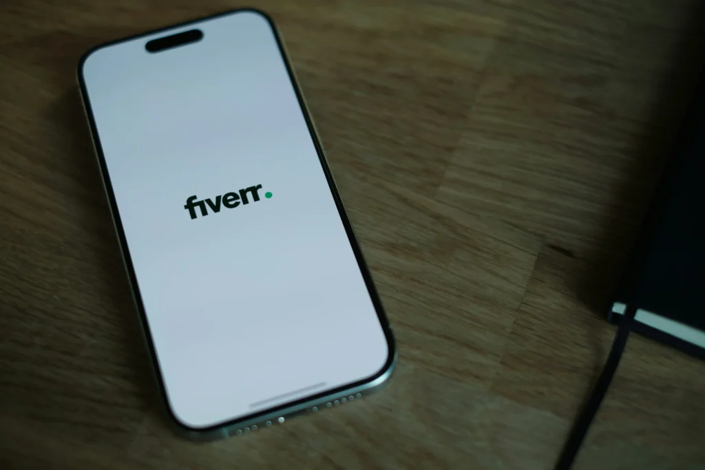 Smartphone displaying Fiverr app logo on screen placed on a wooden desk