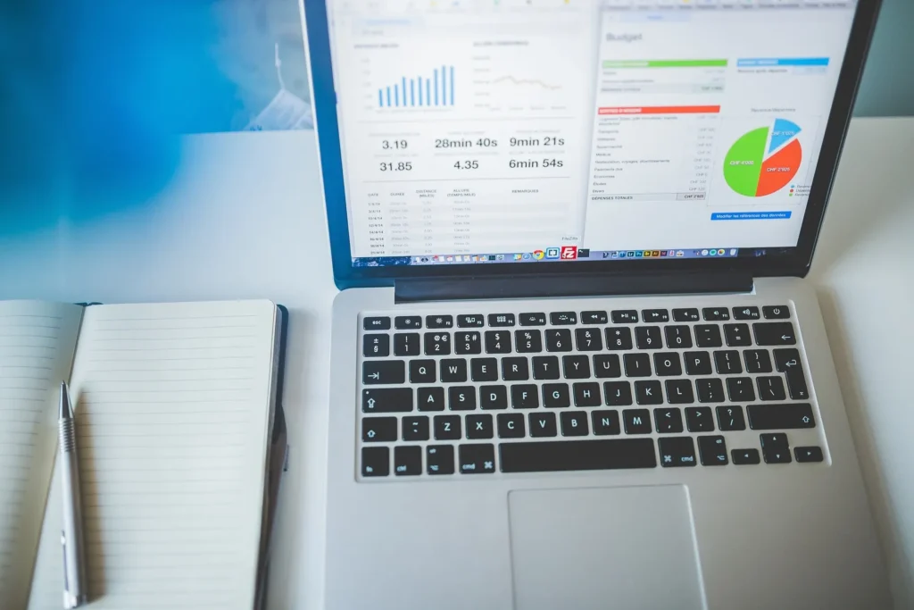 Laptop displaying business analytics dashboard with charts and graphs on a desk next to a notebook and pen